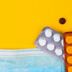 Healthcare items, including a surgical face mask, thermometer, and various pills and capsules, scattered on a yellow surface with the tip of a pill bottle emerging on the bottom right.