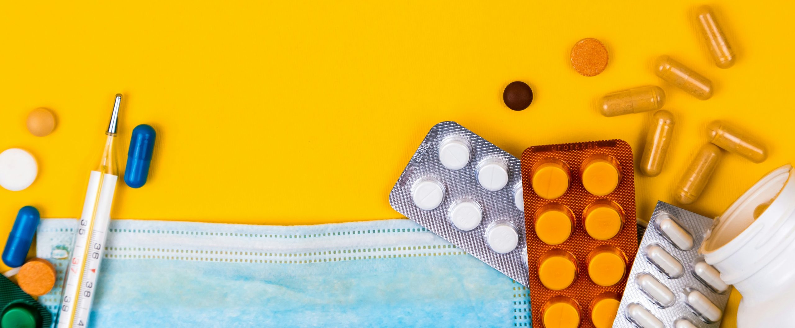 Healthcare items, including a surgical face mask, thermometer, and various pills and capsules, scattered on a yellow surface with the tip of a pill bottle emerging on the bottom right.