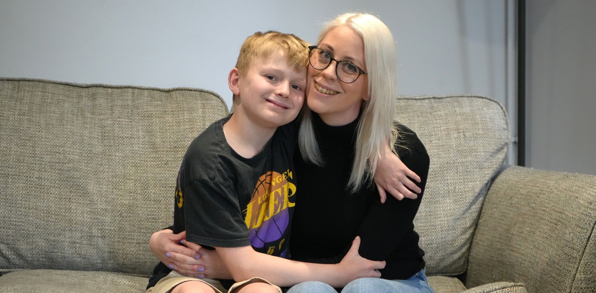 Thomas Farmer and his mother sit on a grey sofa, holding each other and smiling.